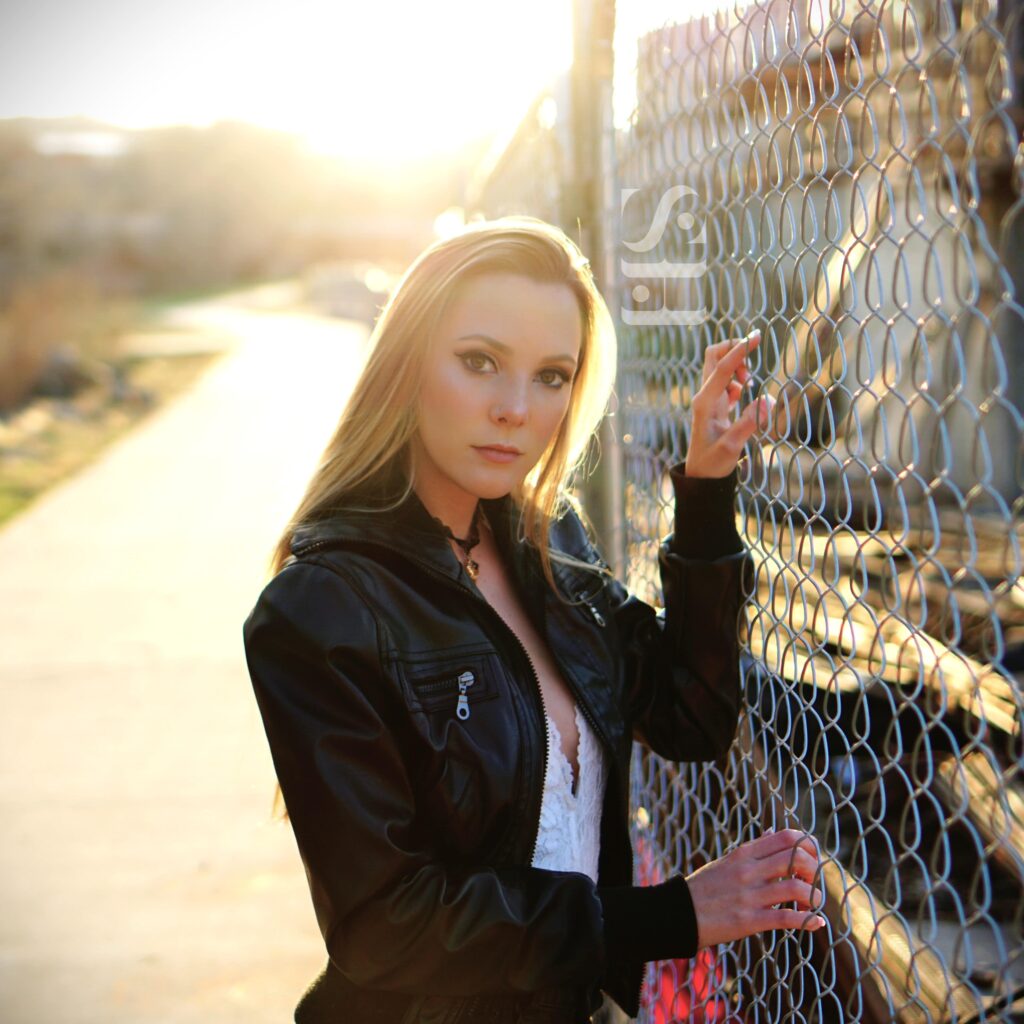 A woman in a black jacket stands by a chain-link fence, gazing at the camera. Sunlight from Longmont creates a warm backdrop as she holds the fence with one hand, captured flawlessly by Pixelhaus Photography.