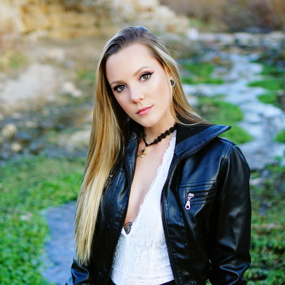 A woman with long blonde hair wearing a black leather jacket and white lace top stands outdoors near a stream surrounded by lush greenery, captured beautifully by Pixelhaus Photography.