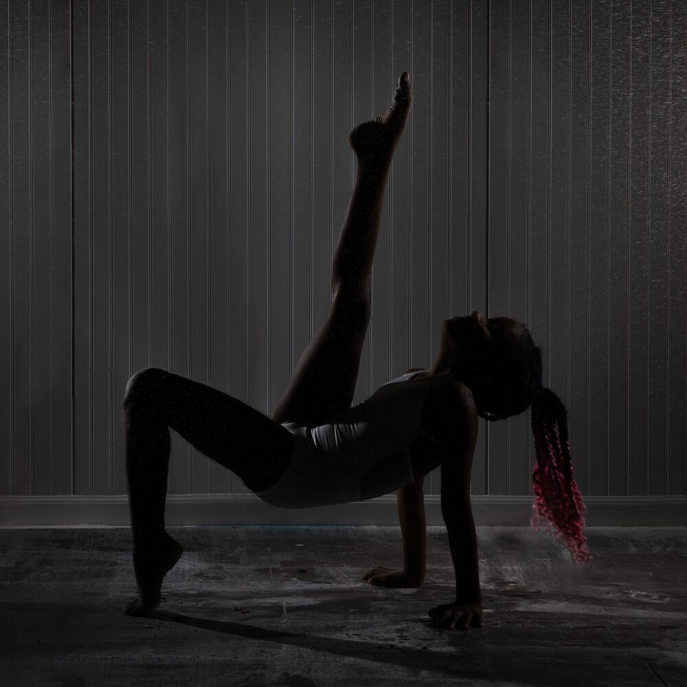 A silhouetted dancer with pink braids performs a one-legged bridge pose against a backlit vertical striped background, captured by Pixelhaus Photography.