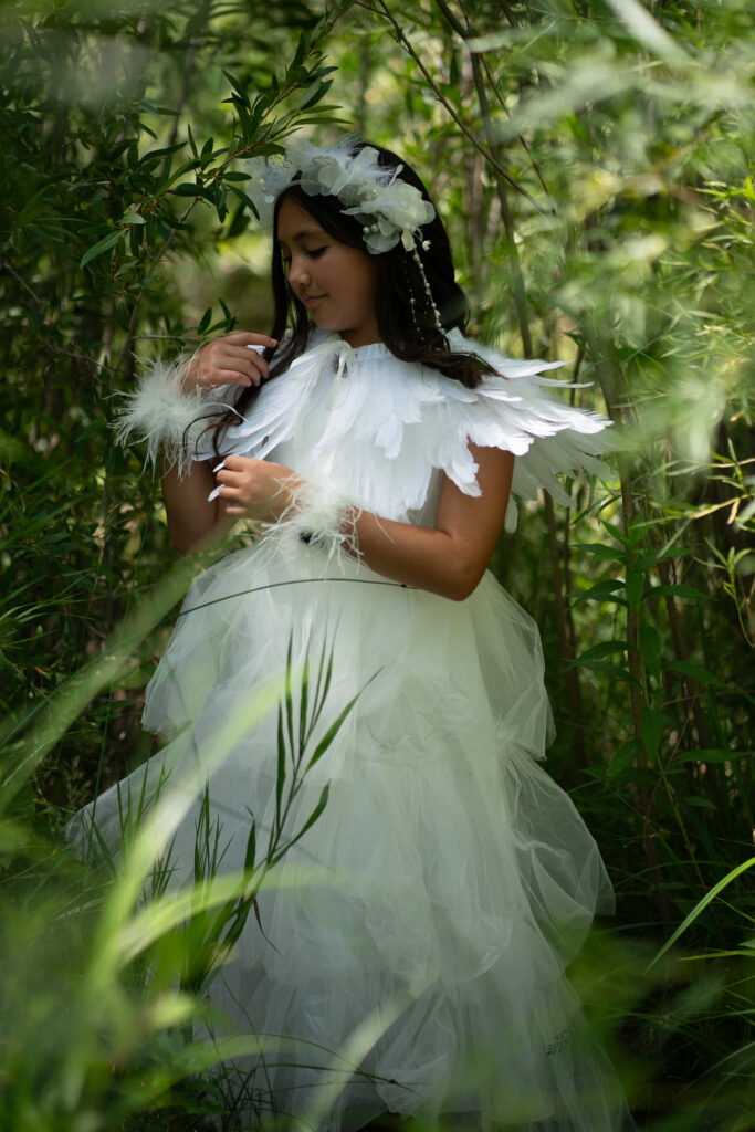 A girl in a white dress and feather accessories stands among green foliage, playing with her hair, beautifully captured by Longmont Photographer.