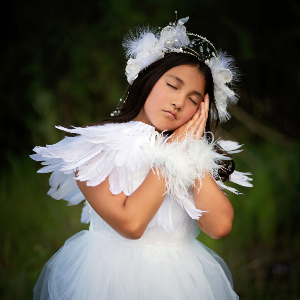A young girl in a white feathered dress and headpiece poses gracefully, her eyes closed and hands gently against her cheek, captured exquisitely by Pixelhaus Photography against a dark green background.