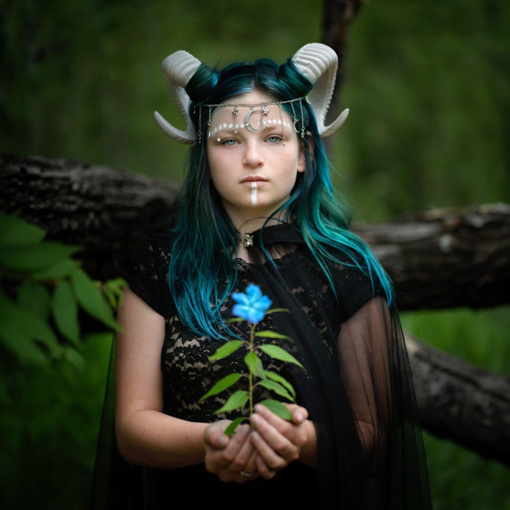 In the lush forest, a person with teal hair and white horns, adorned in a black outfit and necklace, delicately holds a blue flower. Captured by Pixelhaus Photography, this enchanting scene showcases the artistry of a Longmont photographer.