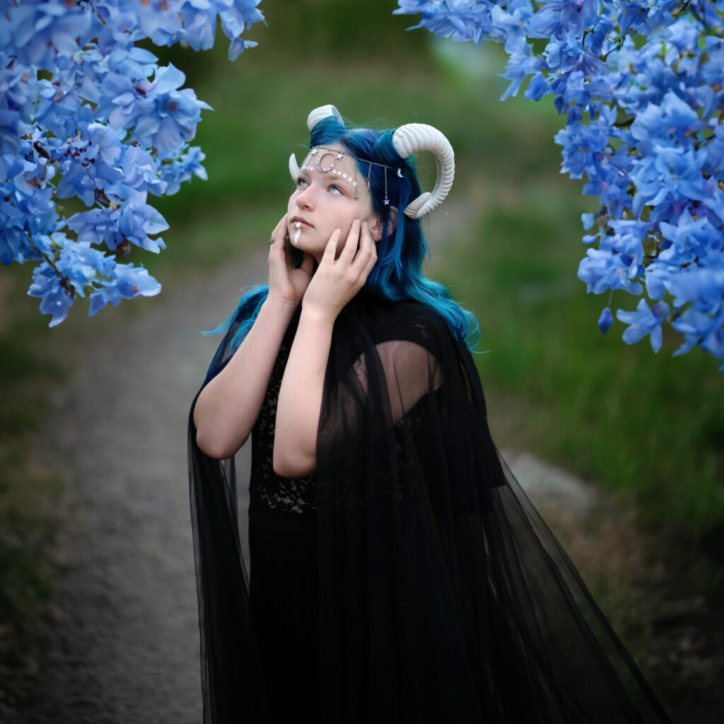 Captured by Pixelhaus Photography, a person with blue hair and a horn headpiece stands on a path surrounded by blue flowers. They wear a black sheer outfit and gaze upward, creating an ethereal scene expertly immortalized by this Longmont photographer.