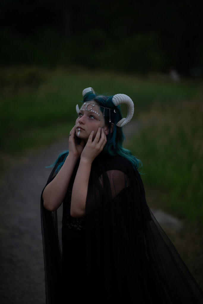 A person with blue hair and fantasy-themed attire, including horns and a dark cloak, stands on a path bordered by grass, gazing contemplatively at the sky—a striking moment captured by Pixelhaus Photography.